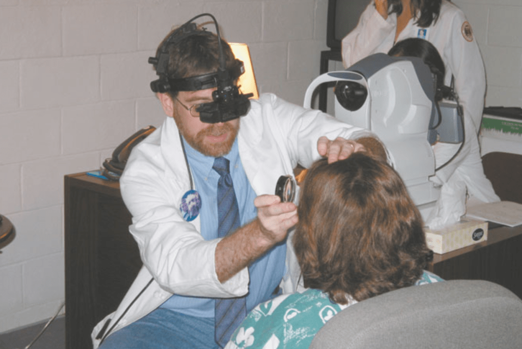 An eye doctor wearing a white coat and a piece of eye equipment on his head holds up a lens to a patients eye to examine it. 