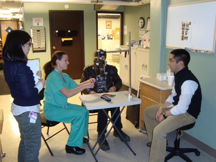 Three student volunteers sit and stand near a visual acuity station to assist a patient. 