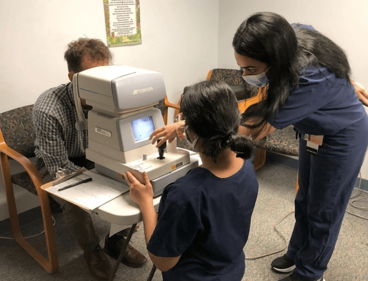 Two medical student volunteers examine a patient using an autorefractor. The patient sits on the opposite side with their face covered, looking into the autorefractor.