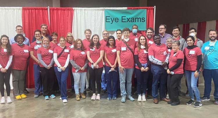 A large group of students, doctors, and other volunteers pose together in a large conference hall under the sign reading "Eye Exams". Almost everyone is wearing a red t-shirt with a heart on it. 